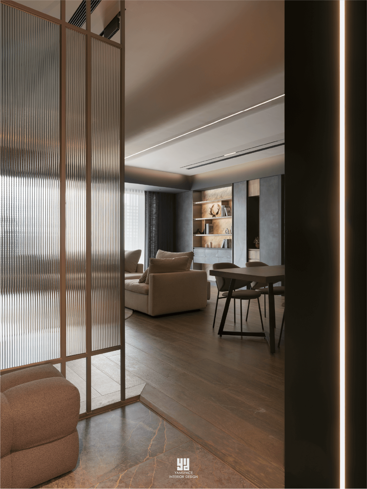 泰御天鑄台中新成屋公領域設計：利用金屬邊框長虹玻璃隔屏作為視覺轉場，透光不透影地串聯客廳與餐廳動線，搭配暗色系櫥櫃、線條感燈帶與深色木地板，展現現代極簡空間深度。